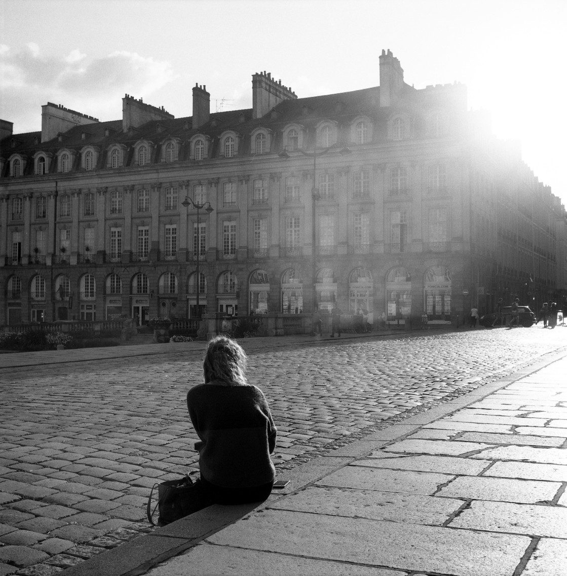 Derrière, photographie argentique noir et blanc d’une silhouette assise dans une place lumineuse