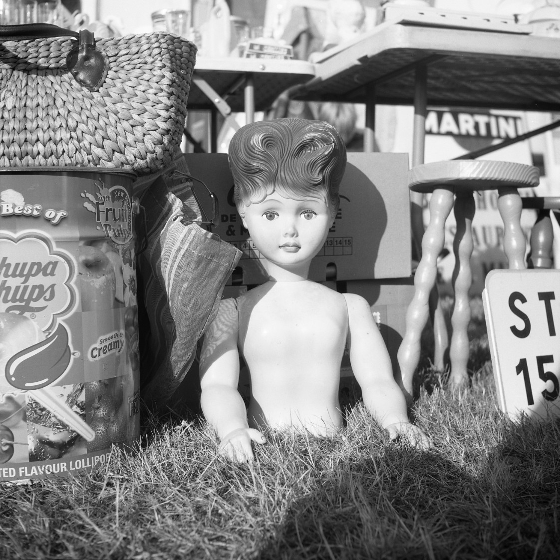 Posée, photographie argentique noir et blanc d’une poupée parmi des objets de brocante